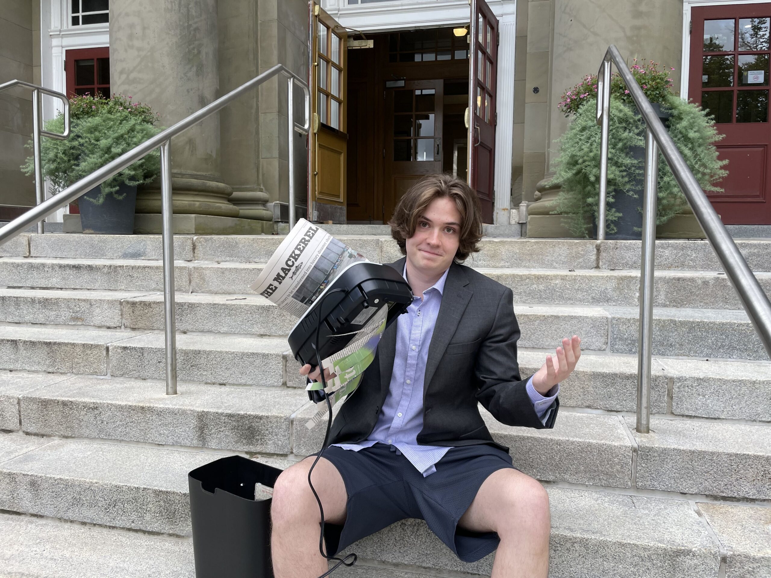 Man in suit jacket and gym shorts shrugging while holding a paper shredder that has an issue of the Dalhousie Gazette halfway through it